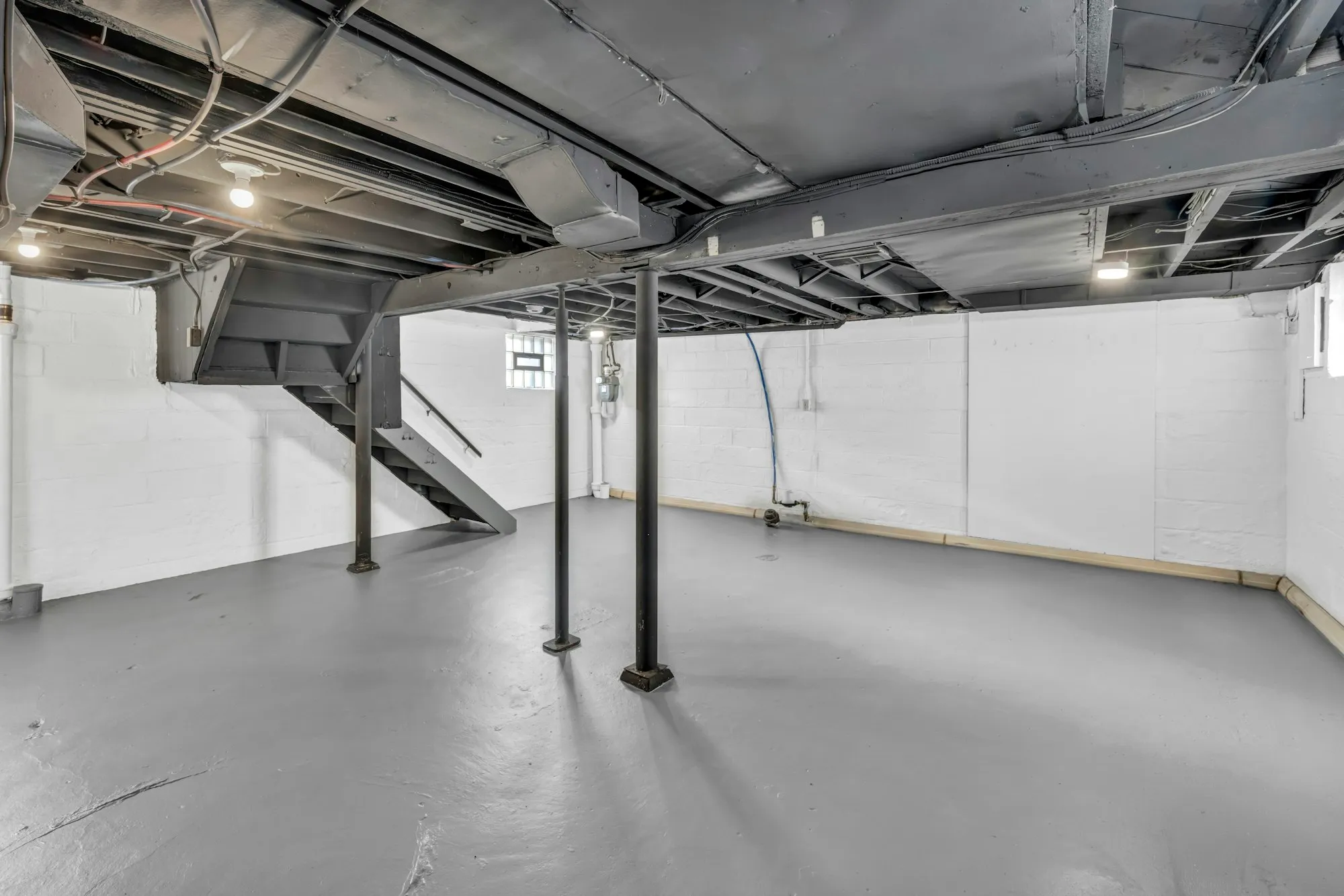 An unfinished basement with exposed floor joists and steel support posts, before development