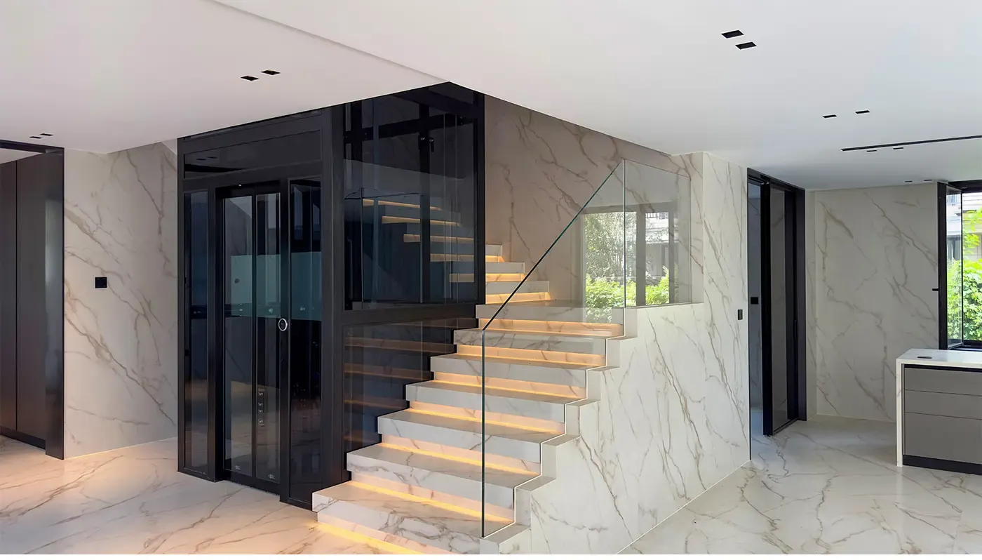 Glass home elevator with a black frame beside a marble staircase